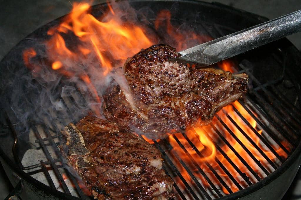 Two ribeye steaks searing over open flame on a charcoal grill